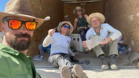 Four paleontologists taking a break inside a storage container, with excavation tools around them and holding a fossil, wearing field hats and sunglasses.