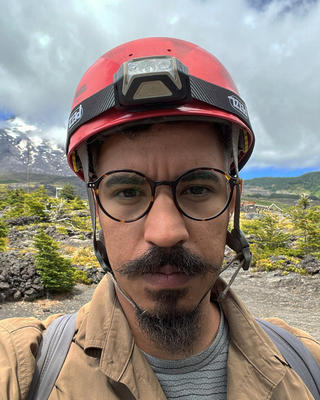 a person looking at the camera, wearing a helmet with a headlamp, outside with greenery and mountains in the background