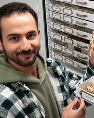 a person smiling at the camera holding a shell in front of drawers of shells