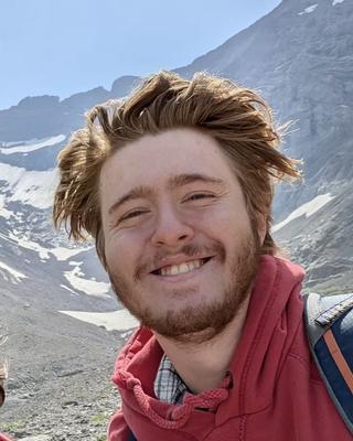 a person smiling at the camera in front of a barren mountain with patches of snow