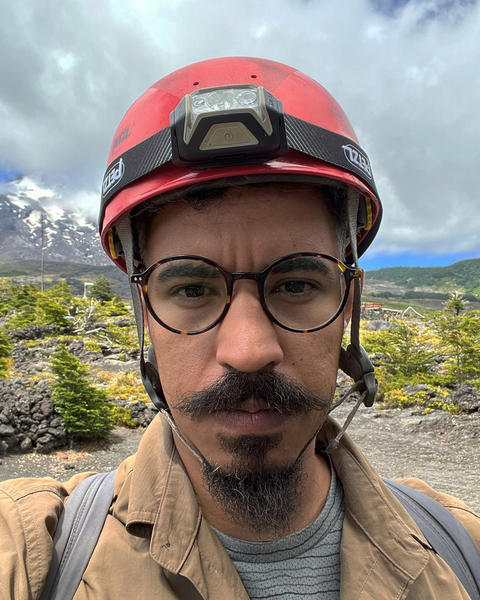 a person looking at the camera, wearing a helmet with a headlamp, outside with greenery and mountains in the background