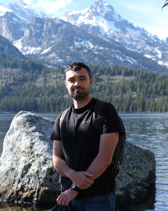 a person smiling at the camera, in front of a lake, forest, and snow-covered mountains