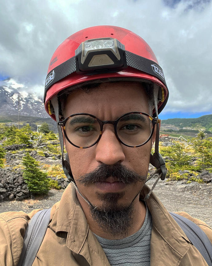a person looking at the camera, wearing a helmet with a headlamp, outside with greenery and mountains in the background
