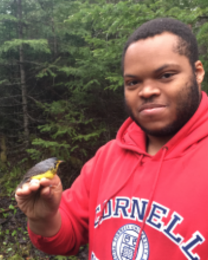 a person looking at the camera, holding a small bird, with green foliage in the background